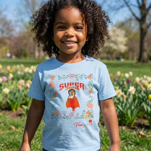 A young child with curly hair stands outdoors in a park wearing a light blue graphic T-shirt with a colorful superhero-style illustration and the words 'SUPER' on the front.