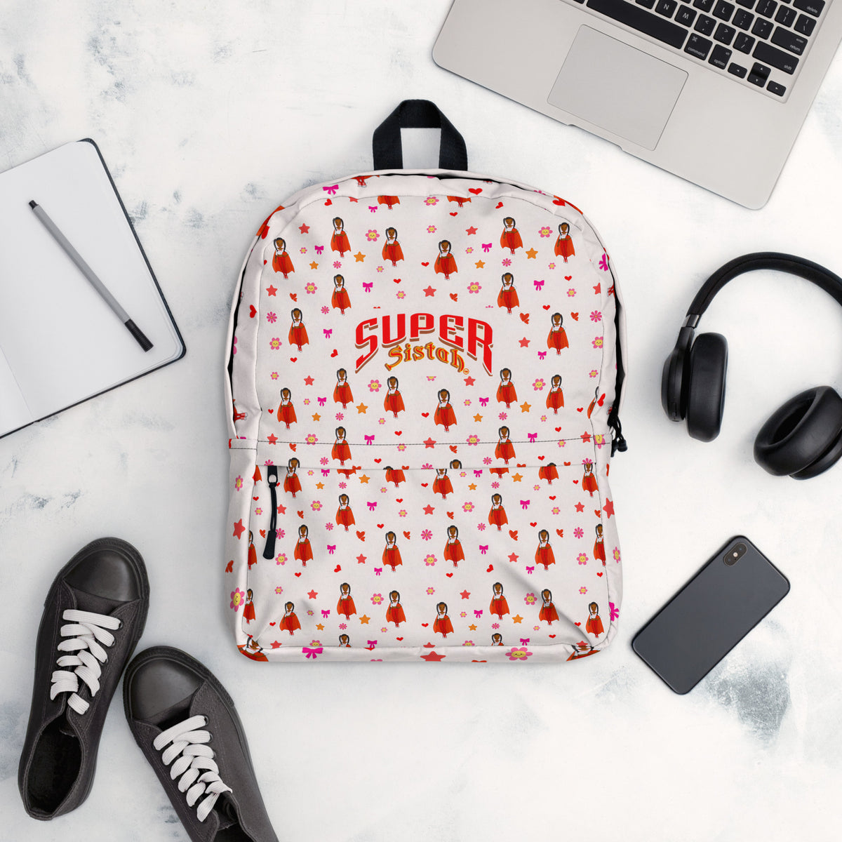 A white backpack with a repeating pattern of cartoon girls in red dresses and the words 'SUPER Sisters' in red and gold, styled in a flat lay with a laptop, headphones, shoes, and a phone on a marble surface.